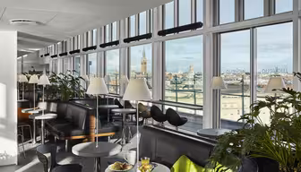 En lys moderne lounge med store vinduer med udsigt over bybilledet. Rummet byder på sorte siddepladser, runde borde med hvide bordplader og grønne planter. På et bord står der en tallerken med mad og et glas appelsinjuice.