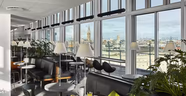 En lys moderne lounge med store vinduer med udsigt over bybilledet. Rummet byder på sorte siddepladser, runde borde med hvide bordplader og grønne planter. På et bord står der en tallerken med mad og et glas appelsinjuice.