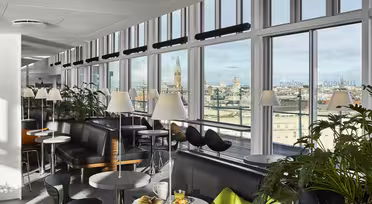 En lys moderne lounge med store vinduer med udsigt over bybilledet. Rummet byder på sorte siddepladser, runde borde med hvide bordplader og grønne planter. På et bord står der en tallerken med mad og et glas appelsinjuice.