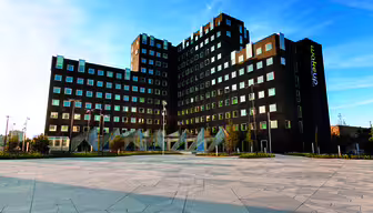 Moderne, kantet sort hotelbygning med flere vinduer mod en klar blå himmel. Forgrunden har en geometrisk, brolagt gårdhave, og et "vågn op"-skilt er synligt på bygningen.