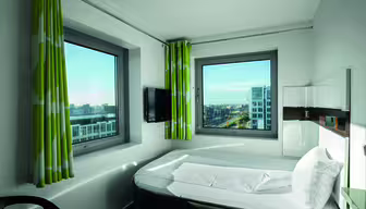 Et moderne hotelværelse med minimalistisk design med en seng, to store vinduer med grønne og hvide gardiner, et væghængt tv og udsigt til bybygninger udenfor. Rummet oplyses af naturligt lys.