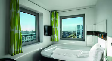Et moderne hotelværelse med minimalistisk design med en seng, to store vinduer med grønne og hvide gardiner, et væghængt tv og udsigt til bybygninger udenfor. Rummet oplyses af naturligt lys.