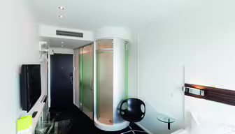 Moderne hotelværelse med en cylindrisk glasbruser nær indgangen. Værelset har en seng, et vægmonteret tv, en sort stol, et sidebord i klart glas og et lille skrivebord med en stol. Hvide vægge og et sort tæppe står i kontrast til minimalistisk indretning.