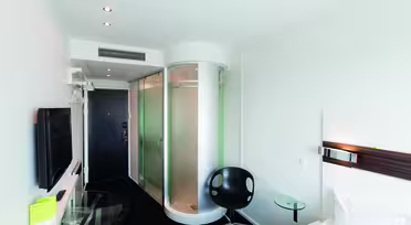 Moderne hotelværelse med en cylindrisk glasbruser nær indgangen. Værelset har en seng, et vægmonteret tv, en sort stol, et sidebord i klart glas og et lille skrivebord med en stol. Hvide vægge og et sort tæppe står i kontrast til minimalistisk indretning.