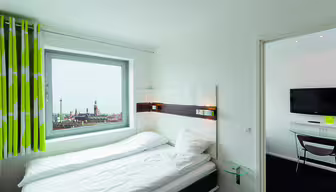 Et rent, lyst hotelværelse har en pænt redt seng med hvidt sengetøj, en væghængt sengegavl med lys og et stort vindue. Udsigten udenfor viser et bybillede med fjerne spir. Grønne og hvide gardiner tilføjer et strejf af farve.