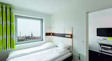 Et rent, lyst hotelværelse har en pænt redt seng med hvidt sengetøj, en væghængt sengegavl med lys og et stort vindue. Udsigten udenfor viser et bybillede med fjerne spir. Grønne og hvide gardiner tilføjer et strejf af farve.