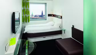 Et moderne hotelværelse med en seng med hvidt sengetøj, en brun sofa, et bord med glasplade og et vægmonteret tv. Et stort vindue giver udsigt til bybilledet med et kirkespir. Rummet har et minimalistisk design med grøn- og hvidstribede gardiner.