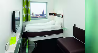 Et moderne hotelværelse med en seng med hvidt sengetøj, en brun sofa, et bord med glasplade og et vægmonteret tv. Et stort vindue giver udsigt til bybilledet med et kirkespir. Rummet har et minimalistisk design med grøn- og hvidstribede gardiner.