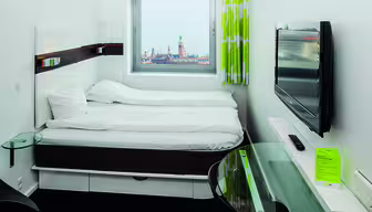 Et moderne hotelværelse med dobbeltseng, væghængt tv og skrivebord i glas. Et vindue giver udsigt til et bybillede og kirketårn. Indretningen omfatter hvidt sengetøj, træaccenter og et grønt gardin med et bladmønster.