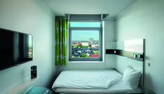 Et lille, moderne hotelværelse med en enkeltseng, vægmonteret tv og et vindue med grønne gardiner, der byder på udsigt over byen. Rummet har et lyst, minimalistisk design med neutrale farver og et glasbord i hjørnet.