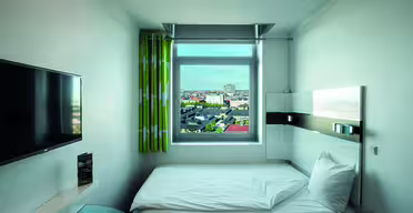Et lille, moderne hotelværelse med en enkeltseng, vægmonteret tv og et vindue med grønne gardiner, der byder på udsigt over byen. Rummet har et lyst, minimalistisk design med neutrale farver og et glasbord i hjørnet.