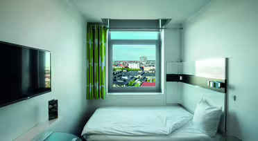 Et lille, moderne hotelværelse med en enkeltseng, vægmonteret tv og et vindue med grønne gardiner, der byder på udsigt over byen. Rummet har et lyst, minimalistisk design med neutrale farver og et glasbord i hjørnet.