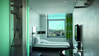 Et moderne hotelværelse med en enkeltseng, tv og et rundt bord. En glasindkapslet bruser er synlig til venstre. Et vindue med udsigt til et bybillede og en delvist overskyet himmel er i baggrunden. Lyse grønne og hvide gardiner hænger ved siden af vinduet.