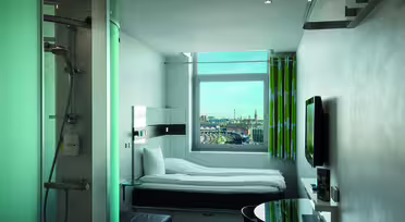 Et moderne hotelværelse med en enkeltseng, tv og et rundt bord. En glasindkapslet bruser er synlig til venstre. Et vindue med udsigt til et bybillede og en delvist overskyet himmel er i baggrunden. Lyse grønne og hvide gardiner hænger ved siden af vinduet.