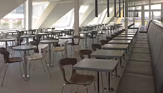 Et moderne, tomt cafeteria med rækker af træstole og firkantede borde. Der er en lang, mørk bænk langs den ene side. Store vinduespartier på den vinklede væg slipper naturligt lys ind. Gulvet er flisebelagt i en lysegrå farve.