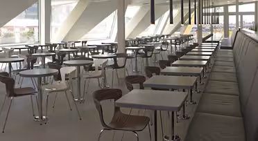 Et moderne, tomt cafeteria med rækker af træstole og firkantede borde. Der er en lang, mørk bænk langs den ene side. Store vinduespartier på den vinklede væg slipper naturligt lys ind. Gulvet er flisebelagt i en lysegrå farve.
