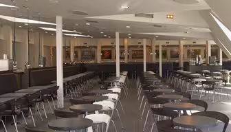 Et stort, moderne cafeteria med rækker af runde borde og stole. Stolene veksler i hvid og mørkebrun. Lokalet er godt oplyst med højt til loftet og har flere maddiske bagerst. Gulvet er flisebelagt i gråt.