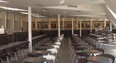 Et stort, moderne cafeteria med rækker af runde borde og stole. Stolene veksler i hvid og mørkebrun. Lokalet er godt oplyst med højt til loftet og har flere maddiske bagerst. Gulvet er flisebelagt i gråt.