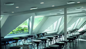 Et rummeligt, moderne cafeteria med rækker af sorte stole og mørke runde borde. Store trekantede vinduer tillader naturligt lys at oplyse rummet og byder på udsigt over frodige grønne områder udenfor. Loftet har indbygget belysning.