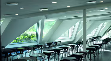 Et rummeligt, moderne cafeteria med rækker af sorte stole og mørke runde borde. Store trekantede vinduer tillader naturligt lys at oplyse rummet og byder på udsigt over frodige grønne områder udenfor. Loftet har indbygget belysning.