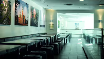 Et elegant, moderne cafeinteriør med en række sorte borde og runde taburetter til venstre. Store, farverige indrammede fotos pryder de hvide vægge. Området er godt oplyst med naturligt lys, der strømmer ind fra højre.