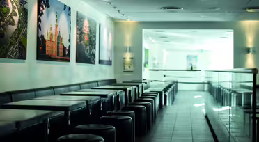 Et elegant, moderne cafeinteriør med en række sorte borde og runde taburetter til venstre. Store, farverige indrammede fotos pryder de hvide vægge. Området er godt oplyst med naturligt lys, der strømmer ind fra højre.