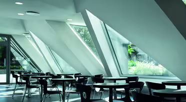 Moderne café med trekantede vinduer og sorte stole omkring runde borde, sat i et lyst rum med naturligt lys, der strømmer ind. Geometrisk arkitektur og grønt synligt gennem glasset skaber en fredfyldt stemning.