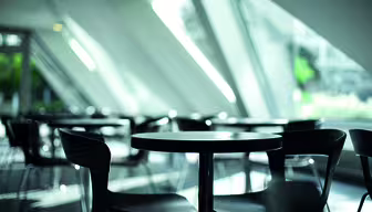Moderne cafeinteriør med slanke sorte stole og runde borde, placeret i nærheden af store, skrå vinduer, der tillader stærkt naturligt lys at strømme ind. Atmosfæren er luftig og minimalistisk, der understreger geometriske arkitektoniske elementer.