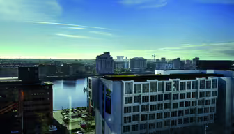 Et bybillede med en havn med moderne bygninger, der omgiver en vandmasse under en klar blå himmel. Scenen omfatter en blanding af arkitektoniske stilarter og et parkeringsområde med biler. Horisonten viser flere bystrukturer.