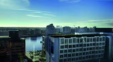 Et bybillede med en havn med moderne bygninger, der omgiver en vandmasse under en klar blå himmel. Scenen omfatter en blanding af arkitektoniske stilarter og et parkeringsområde med biler. Horisonten viser flere bystrukturer.