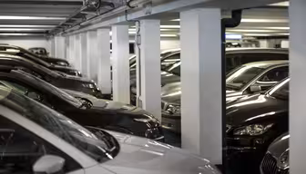 Et svagt oplyst underjordisk parkeringshus fyldt med rækker af parkerede biler. Pladsen er trang med køretøjer i umiddelbar nærhed, og området er understøttet af hvide søjler. Bilerne er for det meste mørkefarvede, med en lysfarvet bil synlig i forgrunden.
