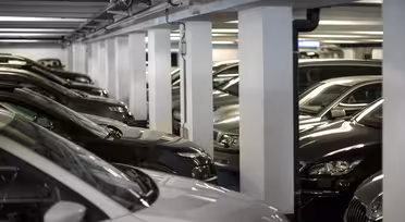 Et svagt oplyst underjordisk parkeringshus fyldt med rækker af parkerede biler. Pladsen er trang med køretøjer i umiddelbar nærhed, og området er understøttet af hvide søjler. Bilerne er for det meste mørkefarvede, med en lysfarvet bil synlig i forgrunden.