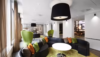 Moderne lounge med buede sorte sofaer og et rundt hvidt bord på et grønt tæppe. Siddeområdet har orange og grå hynder. Høje grønne stole er placeret i nærheden, med store sorte loftslamper, der oplyser rummet. Brune gardiner pryder vinduerne.