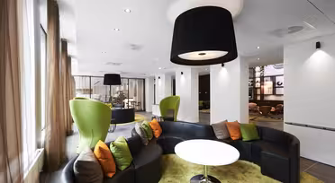 Moderne lounge med buede sorte sofaer og et rundt hvidt bord på et grønt tæppe. Siddeområdet har orange og grå hynder. Høje grønne stole er placeret i nærheden, med store sorte loftslamper, der oplyser rummet. Brune gardiner pryder vinduerne.
