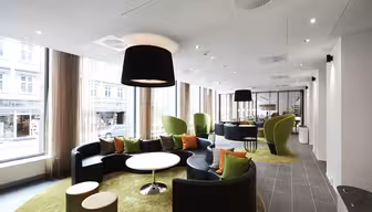 Moderne kontorlounge med cirkulære sorte sofaer, grønne lænestole og runde hvide borde på grønne tæpper. Store vinduer med gennemsigtige gardiner langs siden og lukker naturligt lys ind. Loftslamper og indretning skaber en elegant, åben atmosfære.