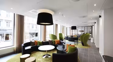 Moderne kontorlounge med cirkulære sorte sofaer, grønne lænestole og runde hvide borde på grønne tæpper. Store vinduer med gennemsigtige gardiner langs siden og lukker naturligt lys ind. Loftslamper og indretning skaber en elegant, åben atmosfære.