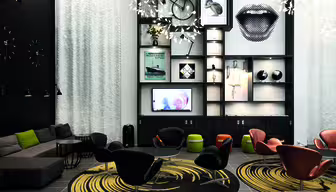 En moderne lounge med eklektisk indretning. Der er et opholdsområde med sorte og orange stole omkring cirkulære tæpper. En væg har hylder med kunst, en cykel og en stor læberillustration. Blød belysning og en sektionssofa fuldender rammerne.