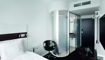 Moderne hotelværelse med en enkeltseng, hvidt sengetøj og en sort stol. En unik cylindrisk glasbruser er synlig nær indgangen. Værelset har minimalistisk indretning med et væghængt tv og et lille glasbord.