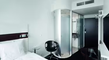 Moderne hotelværelse med en enkeltseng, hvidt sengetøj og en sort stol. En unik cylindrisk glasbruser er synlig nær indgangen. Værelset har minimalistisk indretning med et væghængt tv og et lille glasbord.