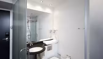 Moderne, minimalistisk badeværelse med hvide vægge og inventar. Har brusekabine af glas, et stort spejl over en sort bordplade, en hvid vask, en vægmonteret hårtørrer og et toilet med en toiletpapirholder. Håndklæder er pænt foldet på disken.