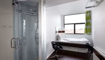 Kompakt moderne værelse med cirkulær glasbruser, enkeltseng ved vinduet, sort drejestol og grønne gardiner. Rummet har hvide vægge og mørkt trægulv, der byder på et minimalistisk design og naturligt lys gennem det store vindue.
