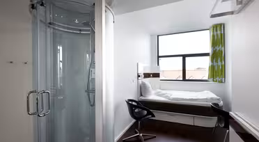 Kompakt moderne værelse med cirkulær glasbruser, enkeltseng ved vinduet, sort drejestol og grønne gardiner. Rummet har hvide vægge og mørkt trægulv, der byder på et minimalistisk design og naturligt lys gennem det store vindue.