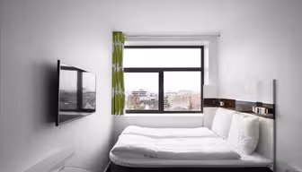 Et lille, moderne hotelværelse med en dobbeltseng mod en hvid væg. Et stort vindue med grønne gardiner tilbyder udsigt over byen. Et fladskærms-tv er monteret på den modsatte væg, og der er et skrivebord med glasplade i nærheden. Rummet er minimalistisk og lyst.