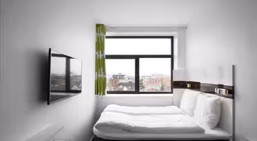 Et lille, moderne hotelværelse med en dobbeltseng mod en hvid væg. Et stort vindue med grønne gardiner tilbyder udsigt over byen. Et fladskærms-tv er monteret på den modsatte væg, og der er et skrivebord med glasplade i nærheden. Rummet er minimalistisk og lyst.