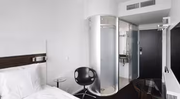 Moderne hotelværelse med en enkeltseng, hvidt sengetøj, en rund sort stol og et lille glasbord. En bruseniche i glas og et kompakt badeværelsesrum er synlige nær indgangen. Indretningen er minimalistisk med hvide vægge og mørkt gulv.