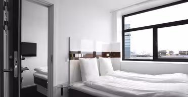Et moderne, minimalistisk hotelværelse med en stor seng dækket af hvidt sengetøj. Der er et fladskærms-tv monteret på væggen, et sidebord i glas og et vindue med udsigt over byens bygninger. Værelset har en ren, lys stemning.