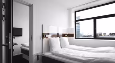 Et moderne, minimalistisk hotelværelse med en stor seng dækket af hvidt sengetøj. Der er et fladskærms-tv monteret på væggen, et sidebord i glas og et vindue med udsigt over byens bygninger. Værelset har en ren, lys stemning.