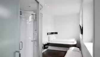 Minimalistisk hotelværelse med hvide vægge og mørkt trægulv. Værelset har en seng, et vægmonteret tv, et lille skrivebord med en stol og en moderne lukket bruser med frostede glasdøre. Stort vindue bringer naturligt lys ind.