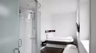 Minimalistisk hotelværelse med hvide vægge og mørkt trægulv. Værelset har en seng, et vægmonteret tv, et lille skrivebord med en stol og en moderne lukket bruser med frostede glasdøre. Stort vindue bringer naturligt lys ind.