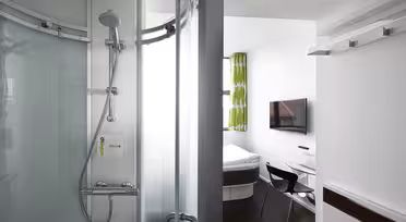 Et kompakt moderne hotelværelse med en buet glasbruser til venstre og et lille soveværelse til højre. Soveværelset har en seng, et væghængt tv, et glasbord og en sort stol. Grønne og hvide mønstrede gardiner dekorerer vinduet.